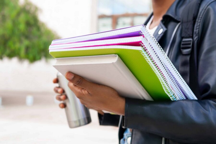 Student wearing backpack carrying stack of spiral-bound notebooks in green, pink, and purple colors along with white textbook while standing outdoors on school campus