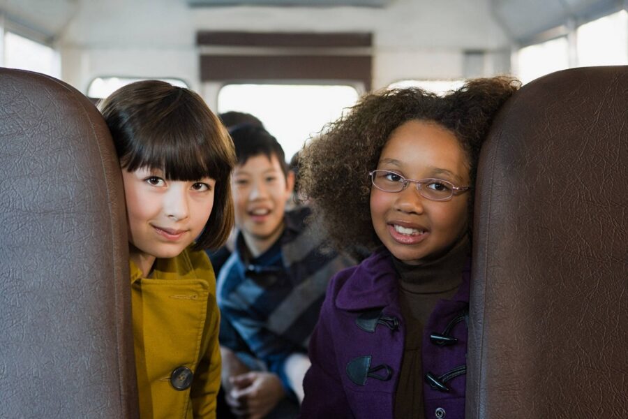 Three diverse elementary school students sitting in brown vinyl bus seats smiling at camera, including girl in yellow shirt and girl with glasses in purple jacket, with other students visible in background
