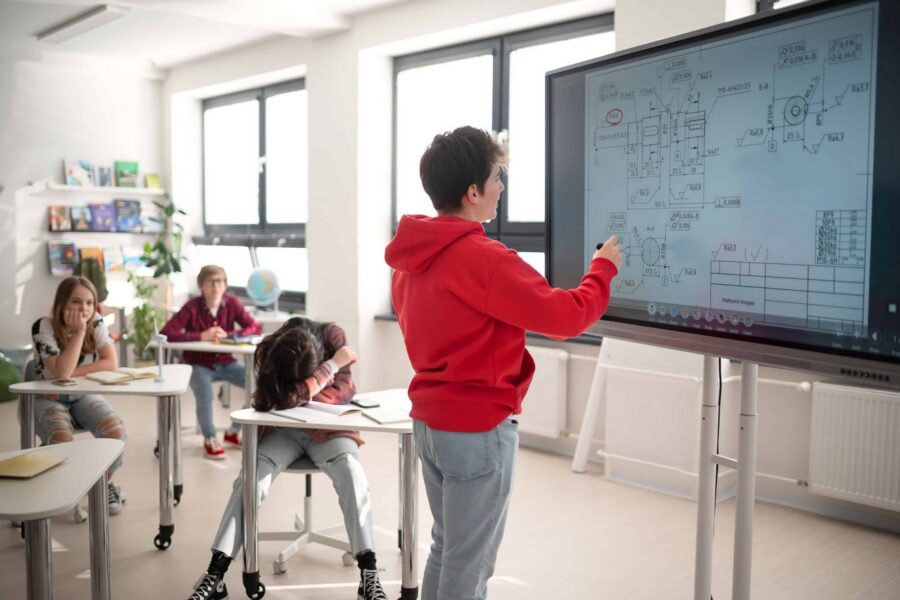 Student in red hoodie presenting technical diagram with mathematical notations and flowcharts on large touchscreen display while classmates observe from desks in bright modern classroom with windows and bookshelves