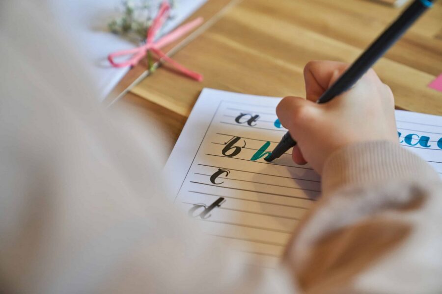 Close-up of child's hand holding black pen practicing lowercase cursive letters 'a', 'b', 'c', 'd' on lined paper worksheet at wooden desk with colorful markers and craft supplies in background