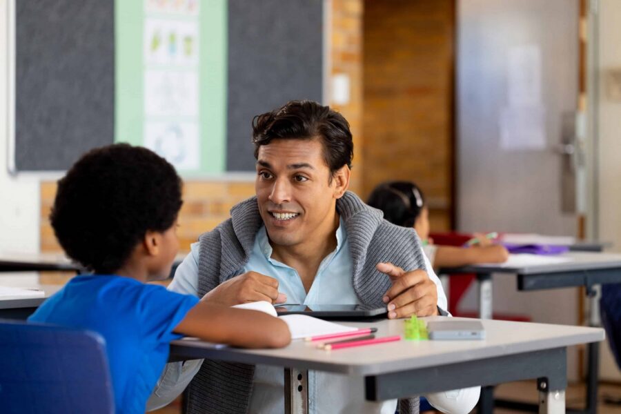 Male teacher in gray cardigan engaging with elementary student during one-on-one instruction at desk with tablet device and learning materials in K-12 classroom setting