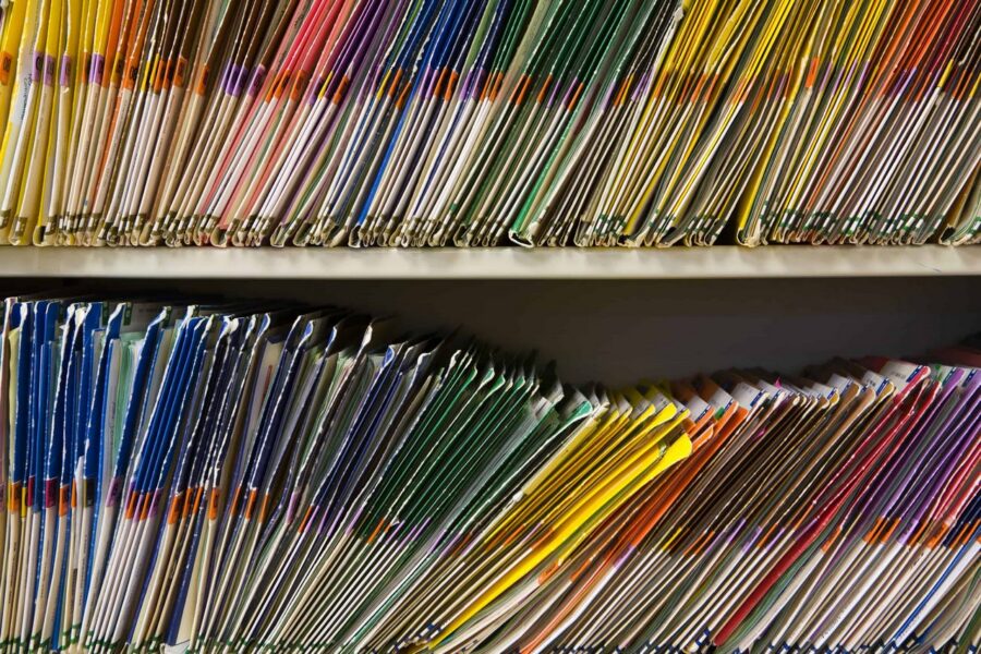 Densely packed color-coded file folders in yellow, orange, red, purple, green, and blue arranged on two-tier shelf system, representing organized document storage and filing system for school district records
