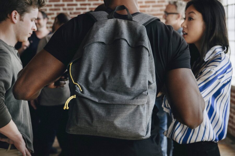 Rear view of person wearing gray backpack with yellow zipper pulls standing among diverse group of participants engaged in discussion during workshop or educational session against exposed brick wall
