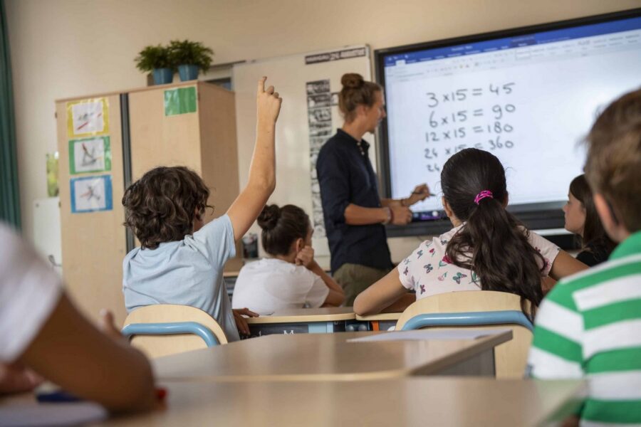Elementary students seated at desks watching teacher present multiplication problems on interactive whiteboard in classroom with one student raising hand to participate