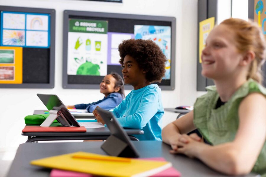 Three diverse elementary school students sitting at desks using digital tablets in classroom with educational displays on monitors behind them, including student in blue shirt focused on tablet with green stand