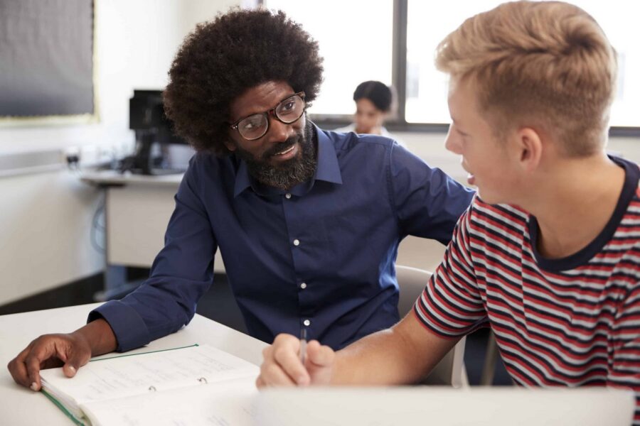 Male teacher with glasses and beard in navy shirt providing one-on-one tutoring to student in striped shirt, both engaged in discussion over open notebook at desk in classroom