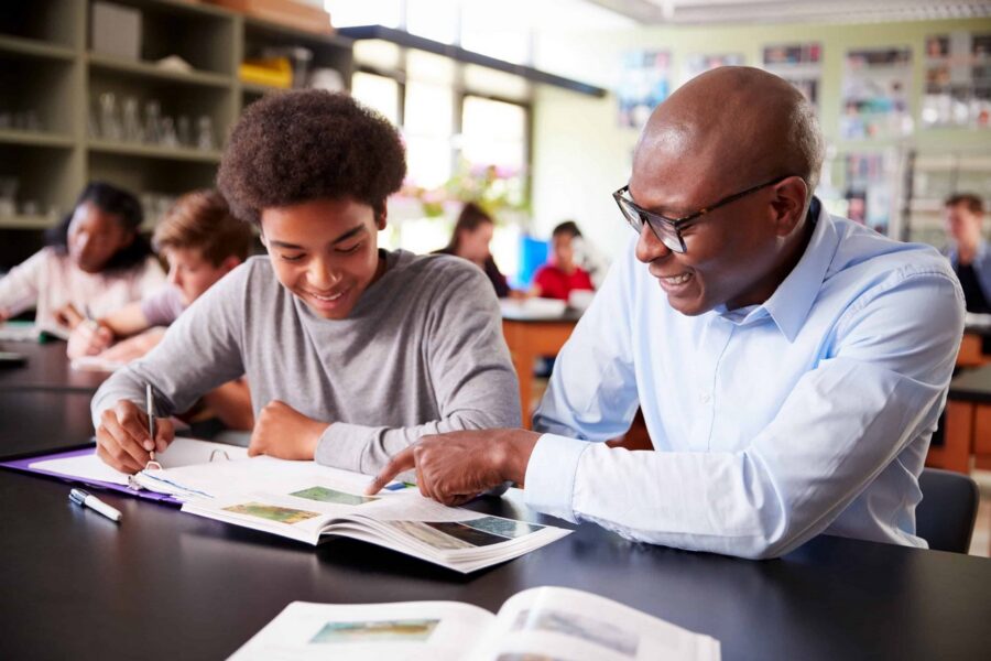 Male teacher in light blue shirt and glasses providing one-on-one tutoring to smiling student in gray shirt, both looking at open textbook together at desk in science classroom with other students working in background