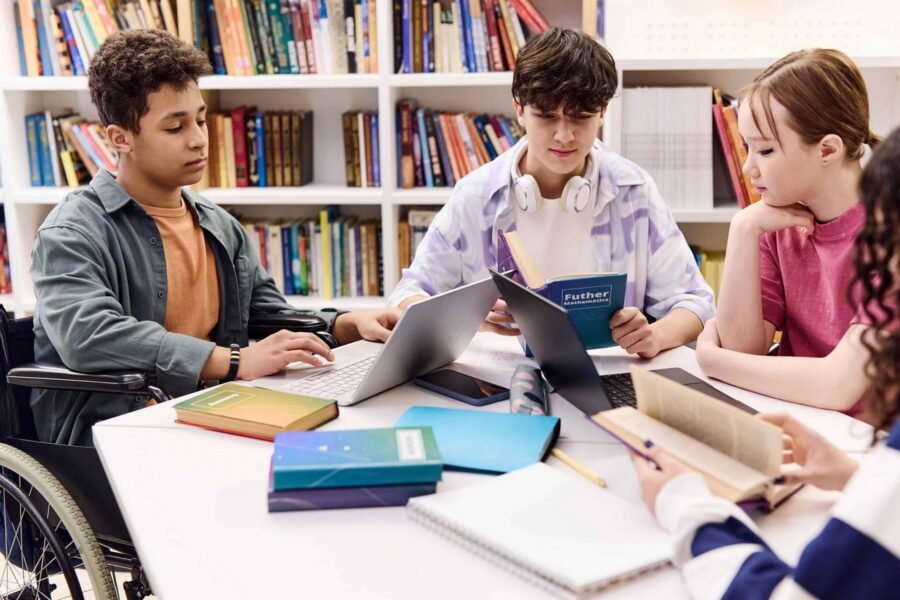 Inclusive group of diverse students collaborating on homework in library with laptops, textbooks including "Further Mathematics" book, and notebooks spread across table in front of bookshelves, featuring student in wheelchair