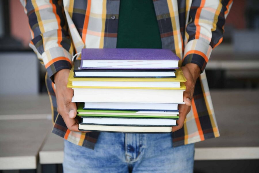 Student in plaid flannel shirt and green t-shirt holding tall stack of colorful textbooks in purple, black, yellow, white, and green covers while standing outdoors on campus