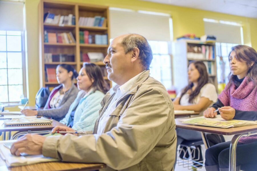 Mature male student in tan jacket sitting at desk with pen and notebook during classroom learning session, with diverse female students visible in background near bookshelves in adult education setting