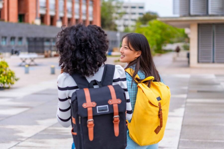Two diverse female students walking together on urban campus, one wearing yellow backpack and plaid shirt smiling at friend wearing striped sweater with black and orange backpack in outdoor school setting