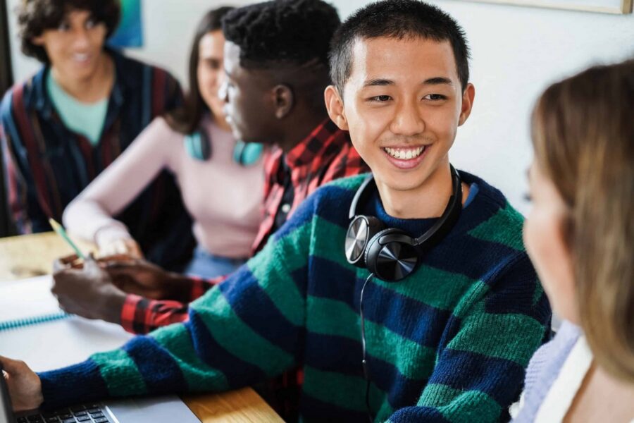 Multiracial group of students studying together at library table with laptops, notebooks, and textbooks, with smiling Asian student wearing striped sweater and headphones in foreground
