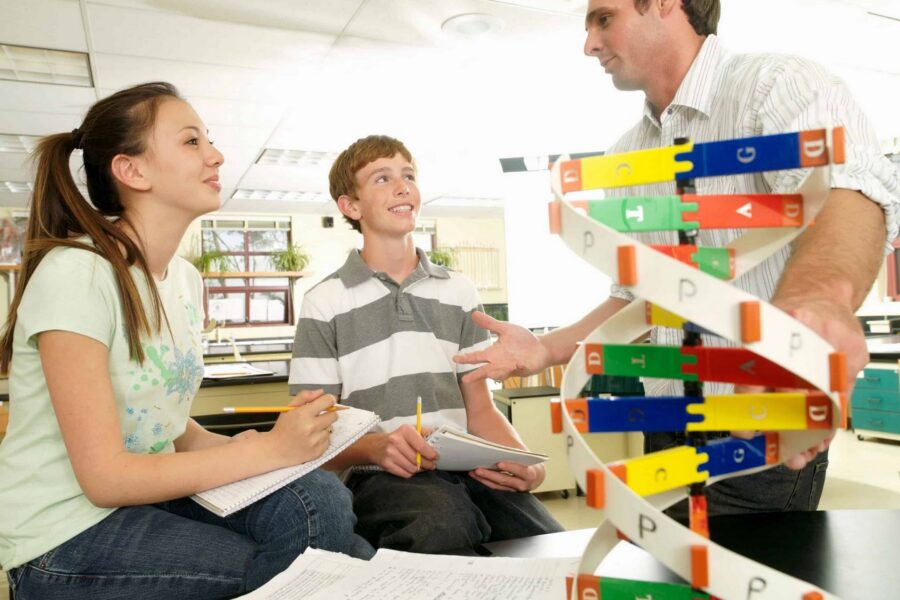 Science teacher in white striped shirt standing with two teenage students observing colorful DNA double helix model made of yellow, blue, green, red, and orange building blocks on desk with notebooks in classroom