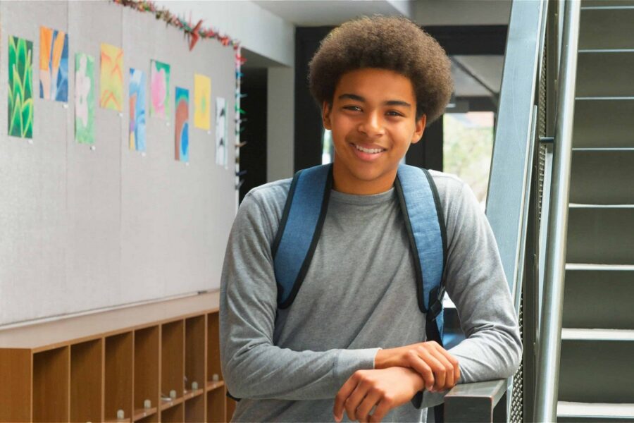 Smiling male high school student wearing blue backpack and gray long-sleeve shirt standing in school hallway with colorful student artwork displayed on wall and wooden lockers in background