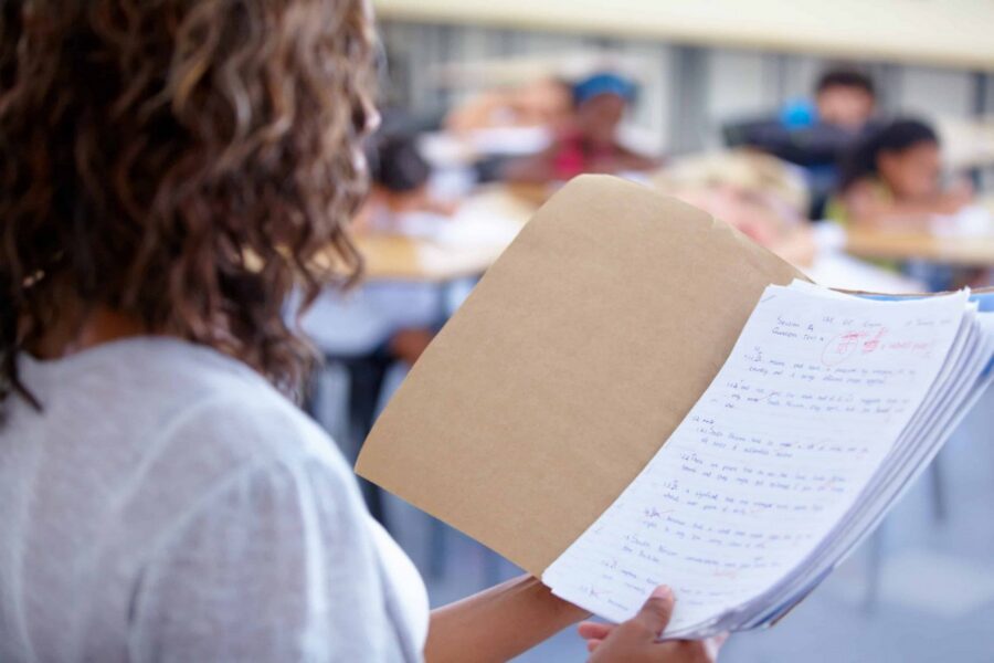 Over-the-shoulder view of student with curly hair in white shirt holding open notebook with handwritten notes and tan folder cover while seated in classroom with blurred students in background