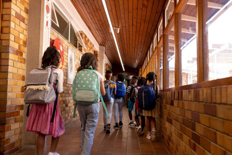 Rear view of diverse group of elementary school children wearing backpacks walking through bright school corridor with wooden ceiling, brick walls, and large windows creating natural lighting