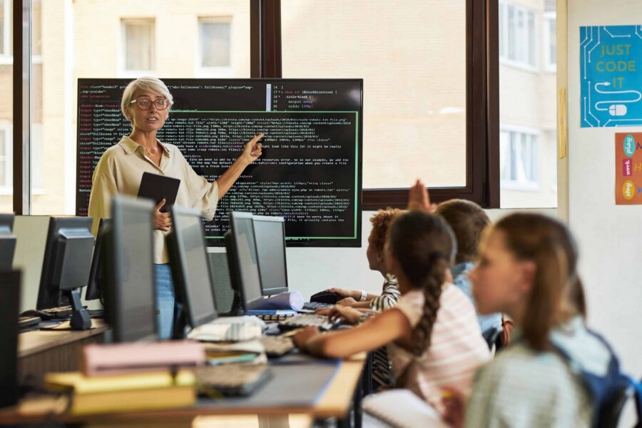 Senior teacher with glasses and yellow shirt holding tablet while teaching coding to diverse elementary students in computer lab, with programming code displayed on large monitor showing HTML and file path examples
