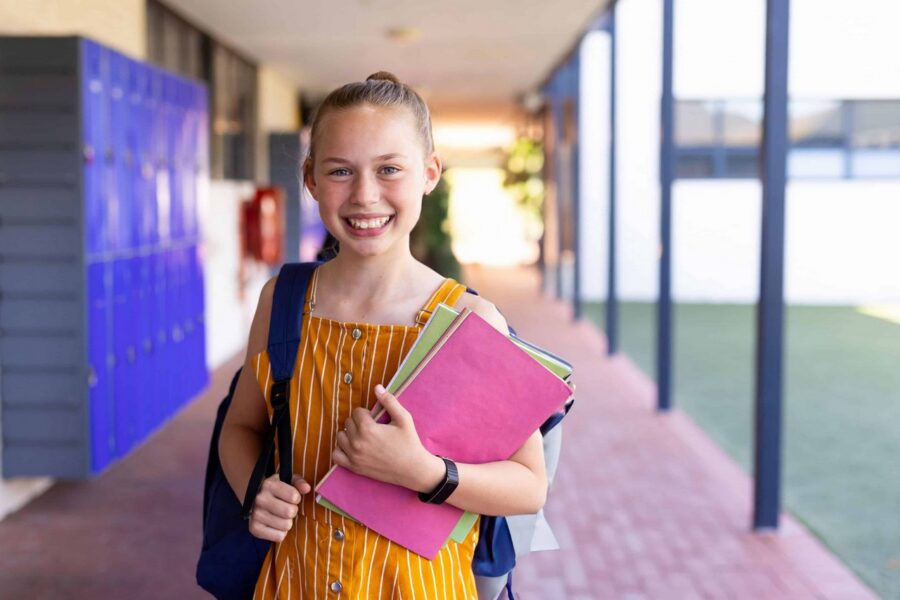 Smiling female student in yellow striped dress wearing navy backpack holding pink and green folders while standing in bright school hallway with blue lockers visible in background