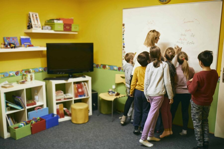 Group of elementary students standing at whiteboard with teacher writing letter A variations while children observe in colorful yellow classroom with TV screen and educational materials on shelves