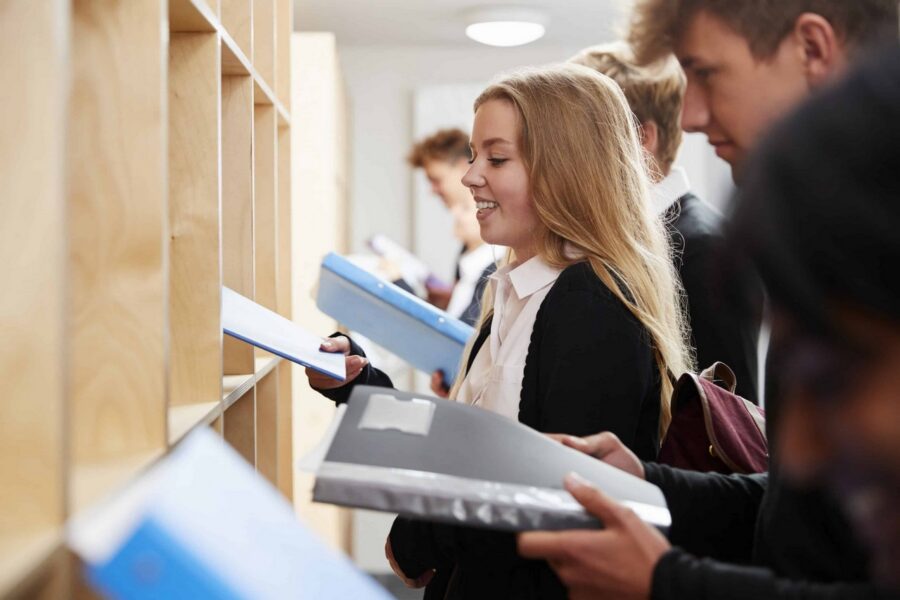 Three teenage students in school uniforms with white collared shirts organizing and putting away blue and gray binders and folders at wooden lockers after class in bright school hallway