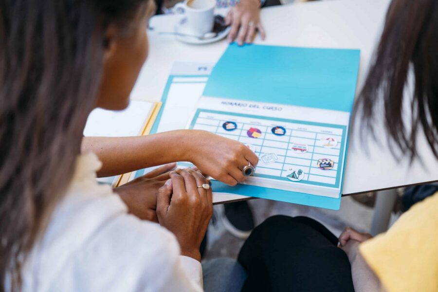 Two students collaborating over a turquoise calendar planner with icons marking different activities, one student pointing at the grid while reviewing educational planning materials at a table